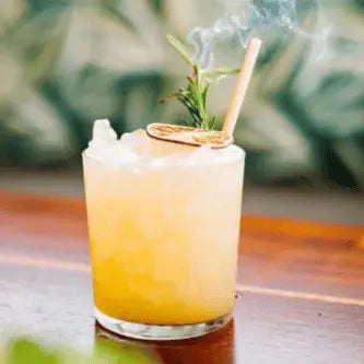 A golden-hued cocktail in a clear glass on a wooden surface, topped with crushed ice, a dried citrus garnish, and a smoking rosemary sprig, featuring a bamboo straw in front of a softly blurred leafy background.