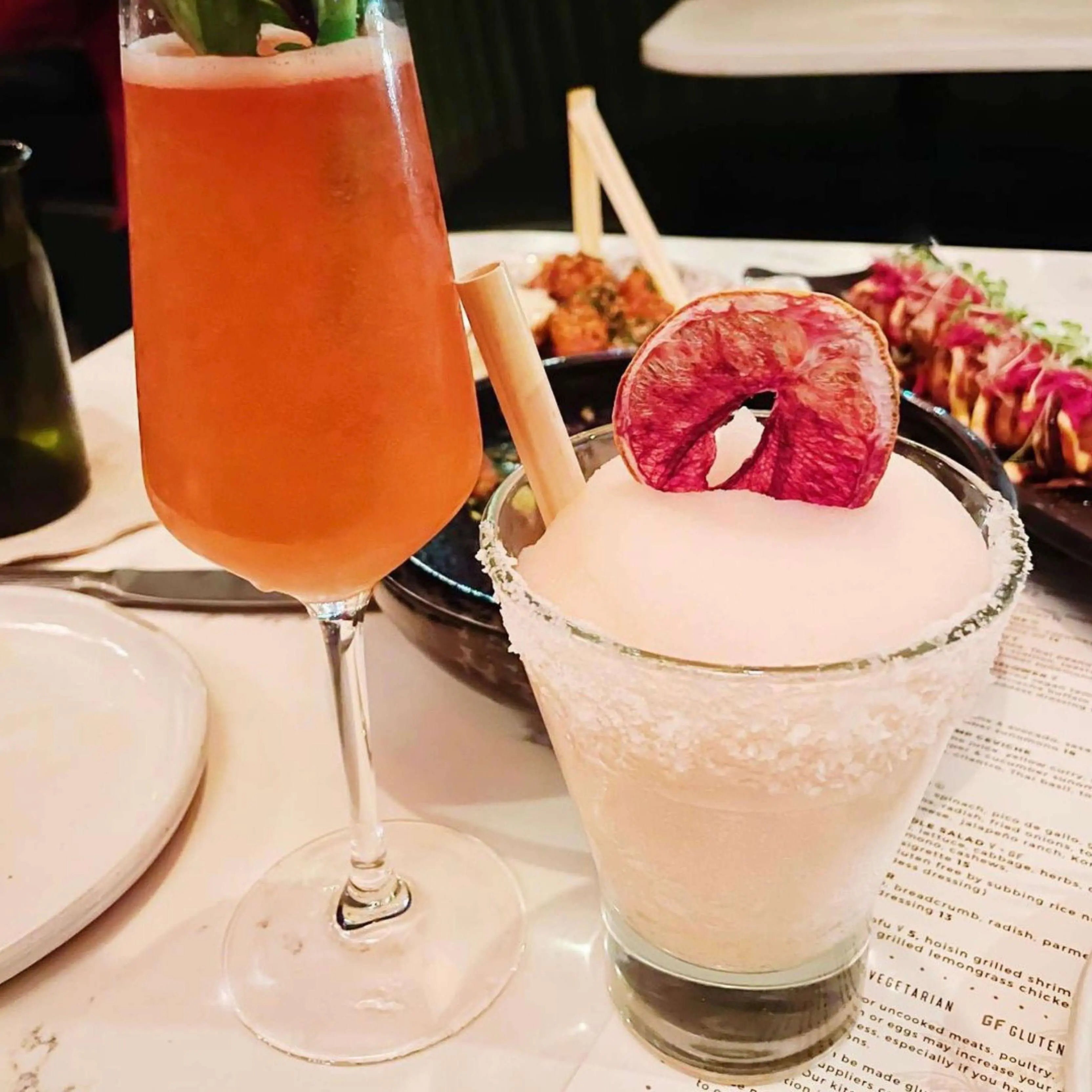 A salted-rim frozen cocktail topped with a dehydrated citrus slice and paired with a flute of pink champagne, both featuring bamboo straws. Small plates of food and another straw are visible in the background.