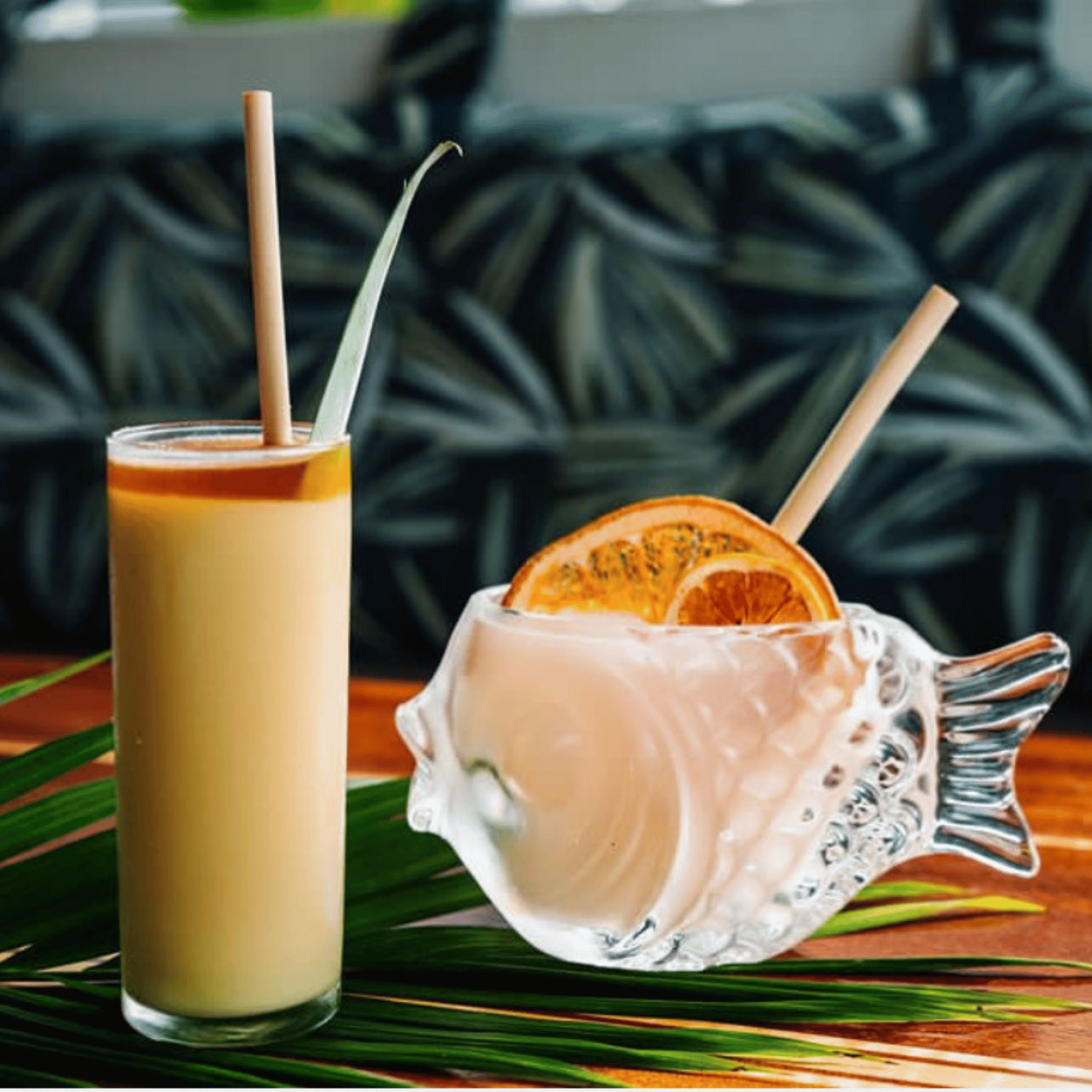 A tall cocktail with a caramel-colored rim and a pineapple leaf garnish is placed beside a clear fish-shaped glass filled with a frozen drink, garnished with dried citrus slices. Both drinks contain bamboo-like straws made from reed stems, which emphasize sustainability. The drinks are set on a wooden surface adorned with tropical leaves, and the blurred background features leaf-patterned upholstery, creating a vibrant and relaxed tropical setting.