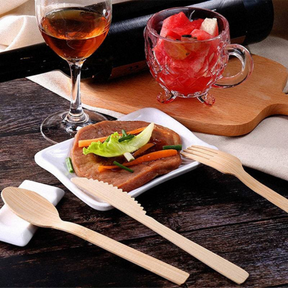 Environmentally-friendly bamboo cutlery set by Holy City Straw Co. displayed on a bamboo cutting board, with a fork, knife, and spoon visible next to slices of fresh broccoli and tomato. Icons below emphasize the utensils' features: Made from Bamboo, 100% Biodegradable, Liquid/Heat Resistant, and Zero Waste