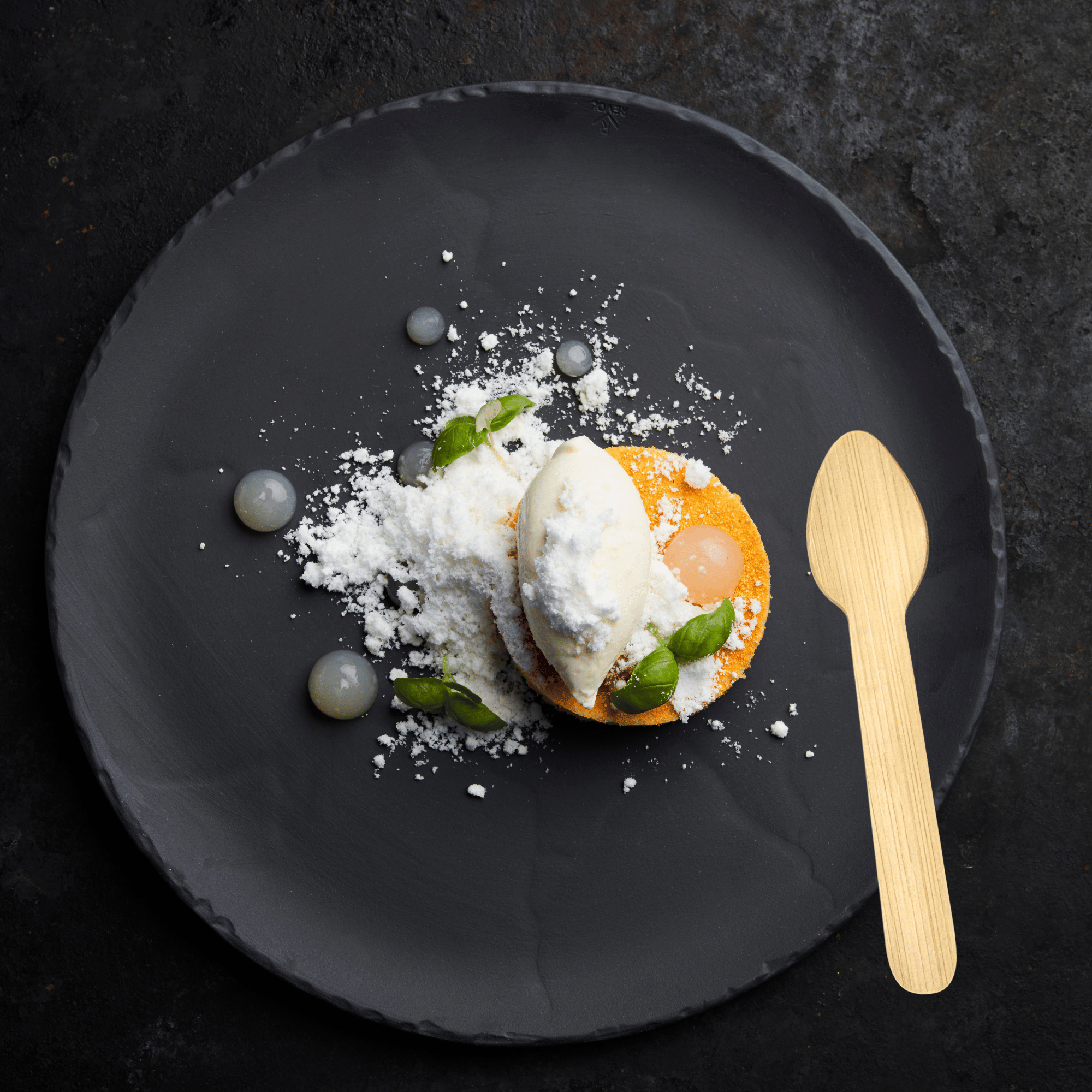 An elegant presentation featuring Holy City Straw Co.'s bamboo dessert spoon on a dark slate plate. The plate holds a deconstructed lemon dessert, with a quenelle of vanilla ice cream atop a crisp round pastry, accompanied by dollops of lemon curd and garnished with fresh basil leaves. Powdered sugar is sprinkled across the arrangement, alongside artistic smears of gray sauce for visual contrast.