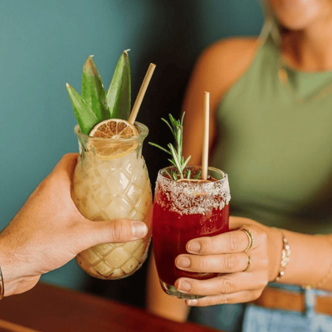 The image shows two people toasting with unique cocktails. On the left, a person holds a cocktail with pineapple garnish and a dried orange slice in a pineapple-shaped glass. The drink on the right, held by a woman, is a red cocktail with a rosemary sprig and salted rim, in a clear glass. Both are enjoying their drinks in a cozy setting with a teal background. The focus is on the drinks and their hands, suggesting a casual, festive atmosphere