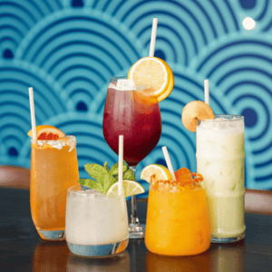 Five colorful cocktails and beverages each with a bamboo fiber straw, set against a modern blue patterned background, highlighting versatility in drink presentation.