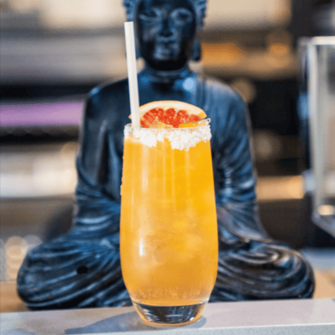 Tall orange cocktail with a bamboo fiber straw and grapefruit garnish placed in front of a dark Buddha statue, capturing a serene, upscale bar environment.