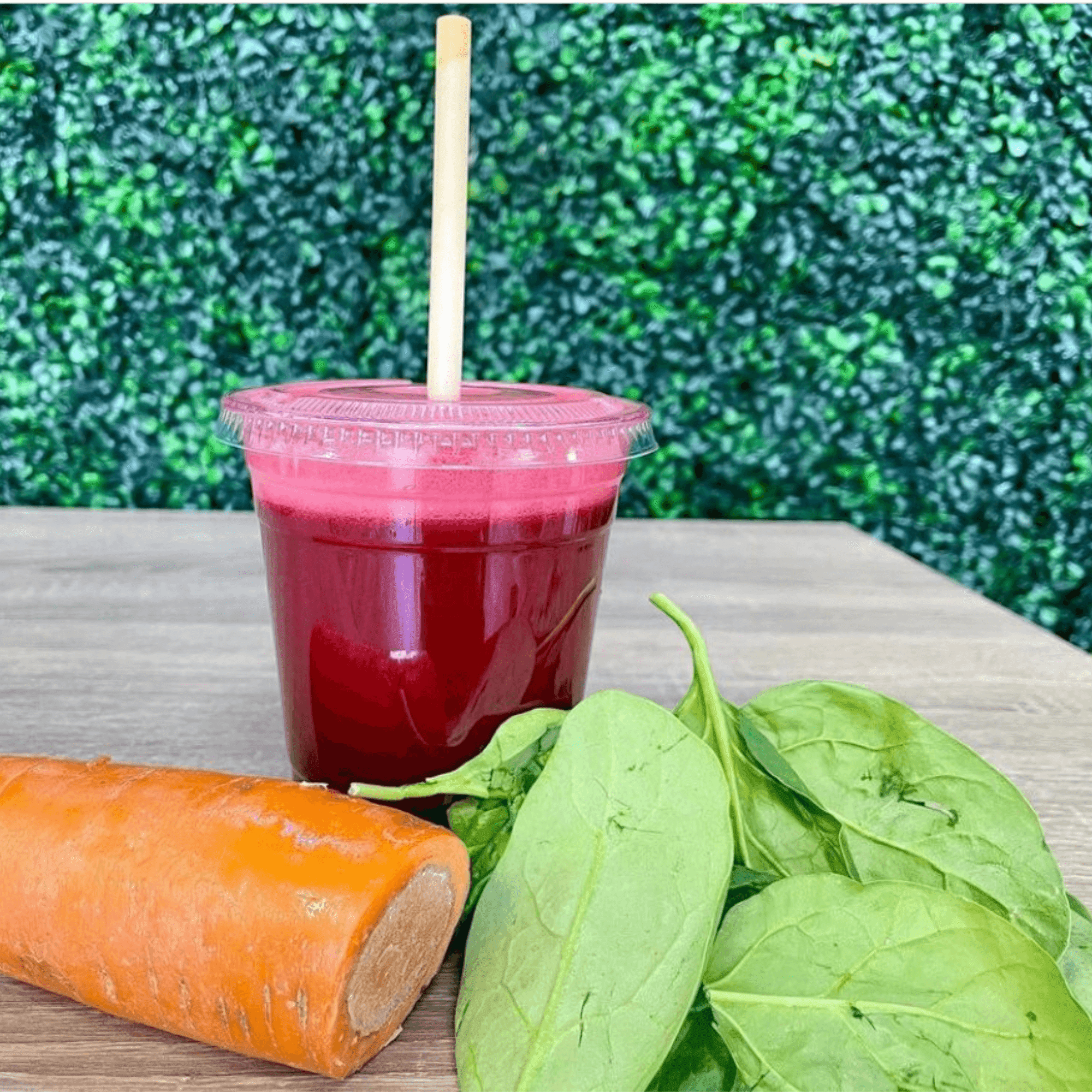 A beet-infused juice is served in a clear cup with a biodegradable straw made from reed stems. The cup is placed on a wooden table next to a raw carrot and fresh spinach leaves. The background is a lush green wall, creating a natural and refreshing atmosphere that emphasizes the juice's healthy and organic ingredients. The straw's sustainable material highlights an eco-friendly and health-conscious approach.