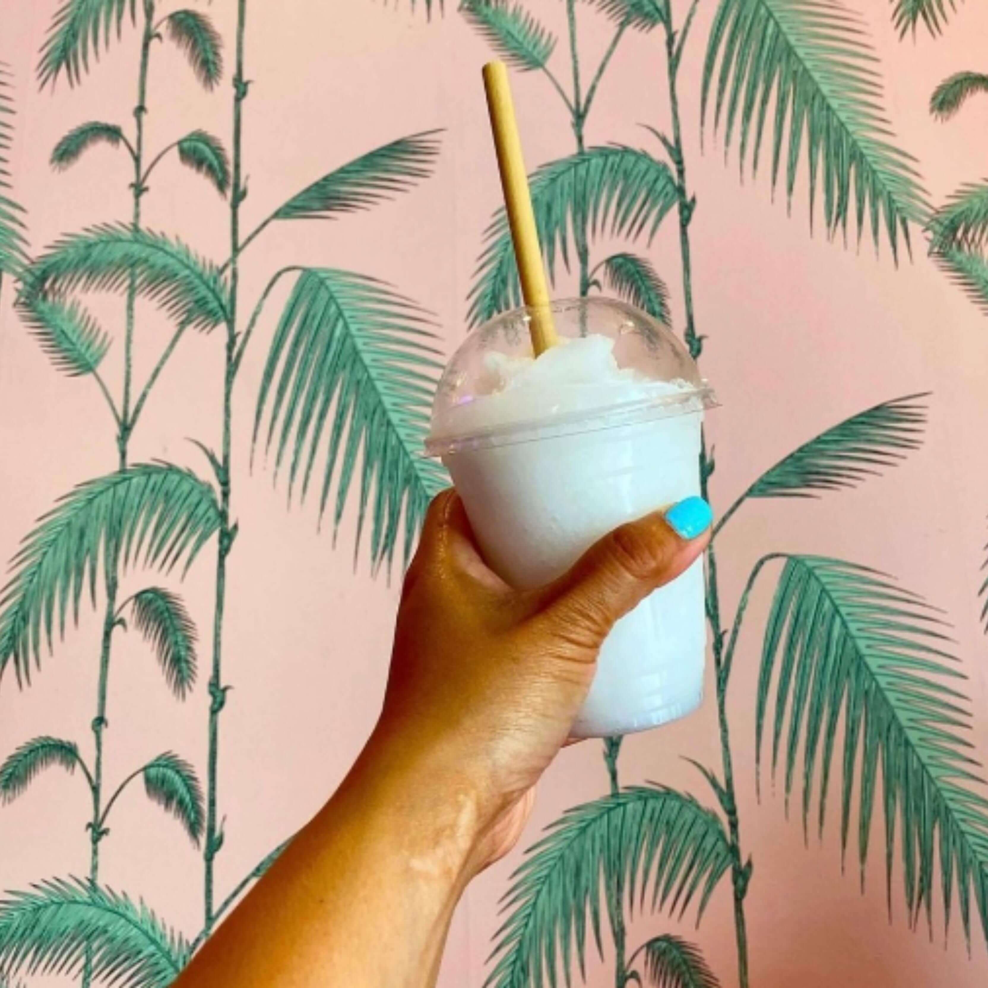 A hand with bright blue nail polish is holding an iced lemonade drink in a clear cup with a dome lid. A bamboo-like straw made from reed stems is inserted through the lid. The background features a pale pink wall with green palm tree designs, giving a tropical and refreshing feel to the scene. The eco-friendly straw and palm backdrop emphasize a playful, sustainable aesthetic.