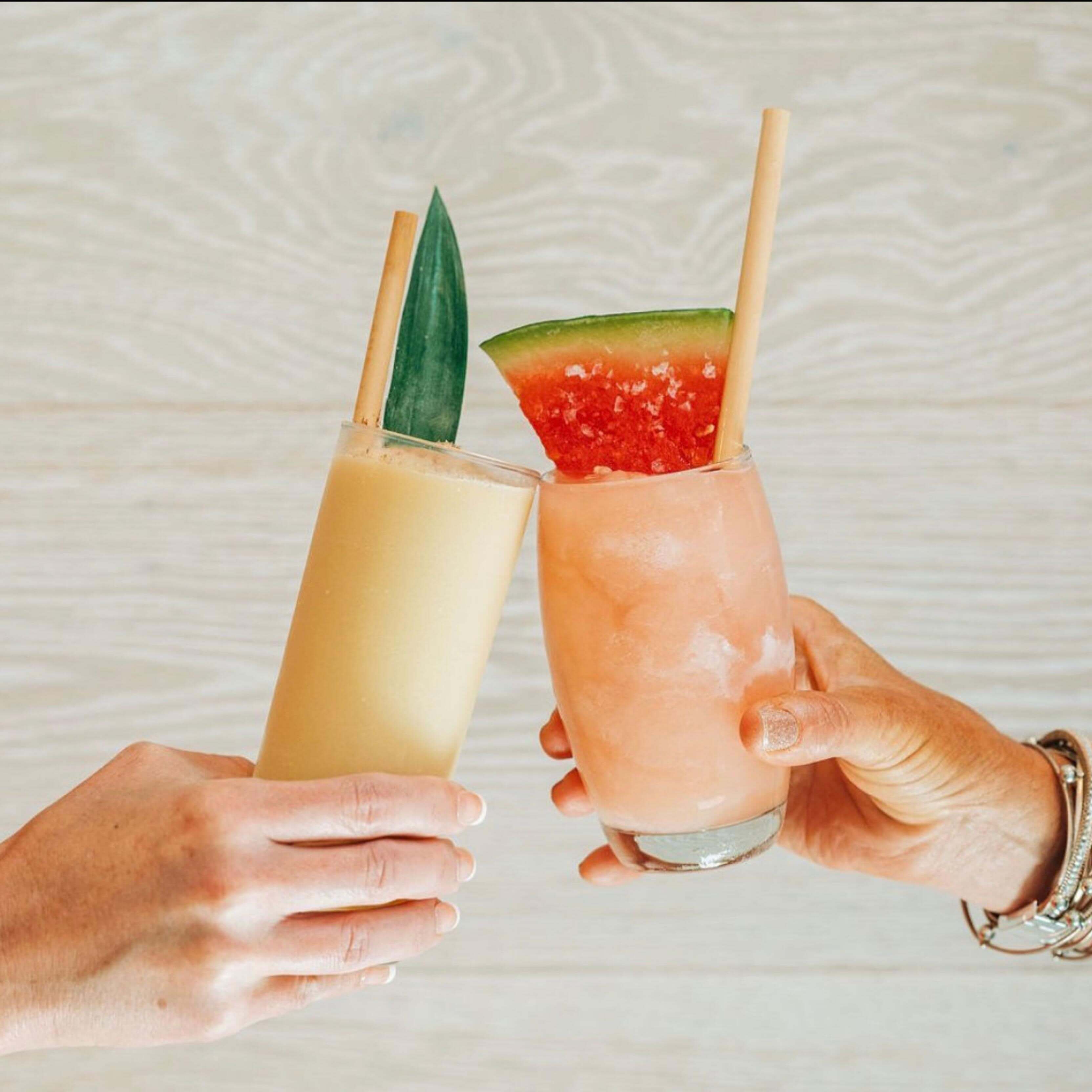 Two people are toasting with fruit cocktails in clear glasses. One cocktail is garnished with a pineapple leaf, while the other has a slice of watermelon. Both drinks feature bamboo-like straws made from reed stems, emphasizing their eco-friendly nature. The background is a light wooden surface, highlighting the colorful drinks and their sustainable presentation.