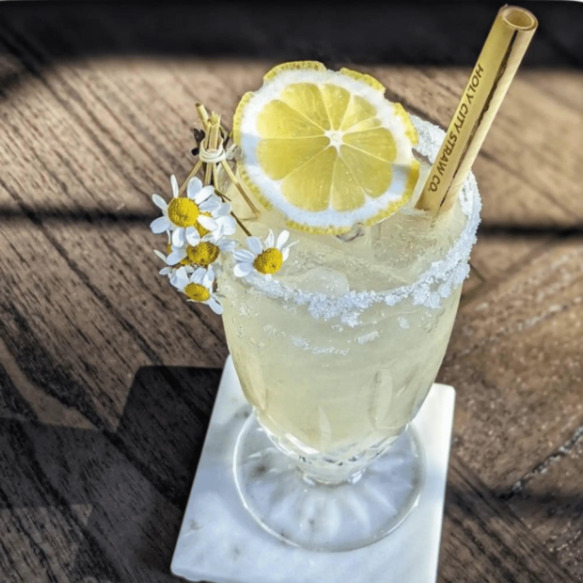 A tall lemon-yellow cocktail in an ornate glass rimmed with sugar, garnished with a fresh lemon slice and small daisies. A bamboo straw printed with ‘Holy City Straw Co.’ stands in the icy drink on a wooden table