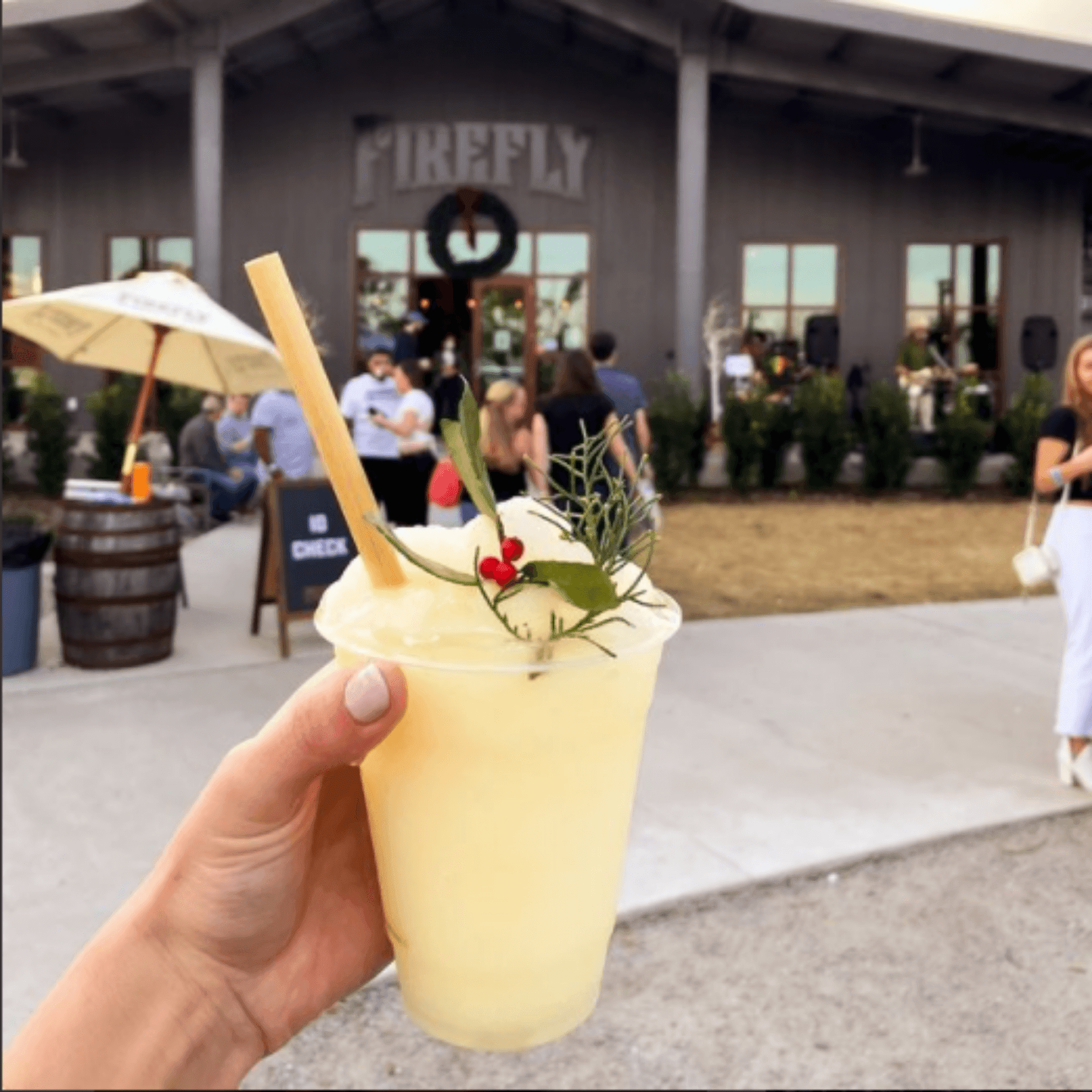 A hand is holding a clear cup filled with frozen lemonade, garnished with sprigs of greenery, berries, and a bamboo-like straw made from reed stems. The setting is outdoors, in front of a venue named 