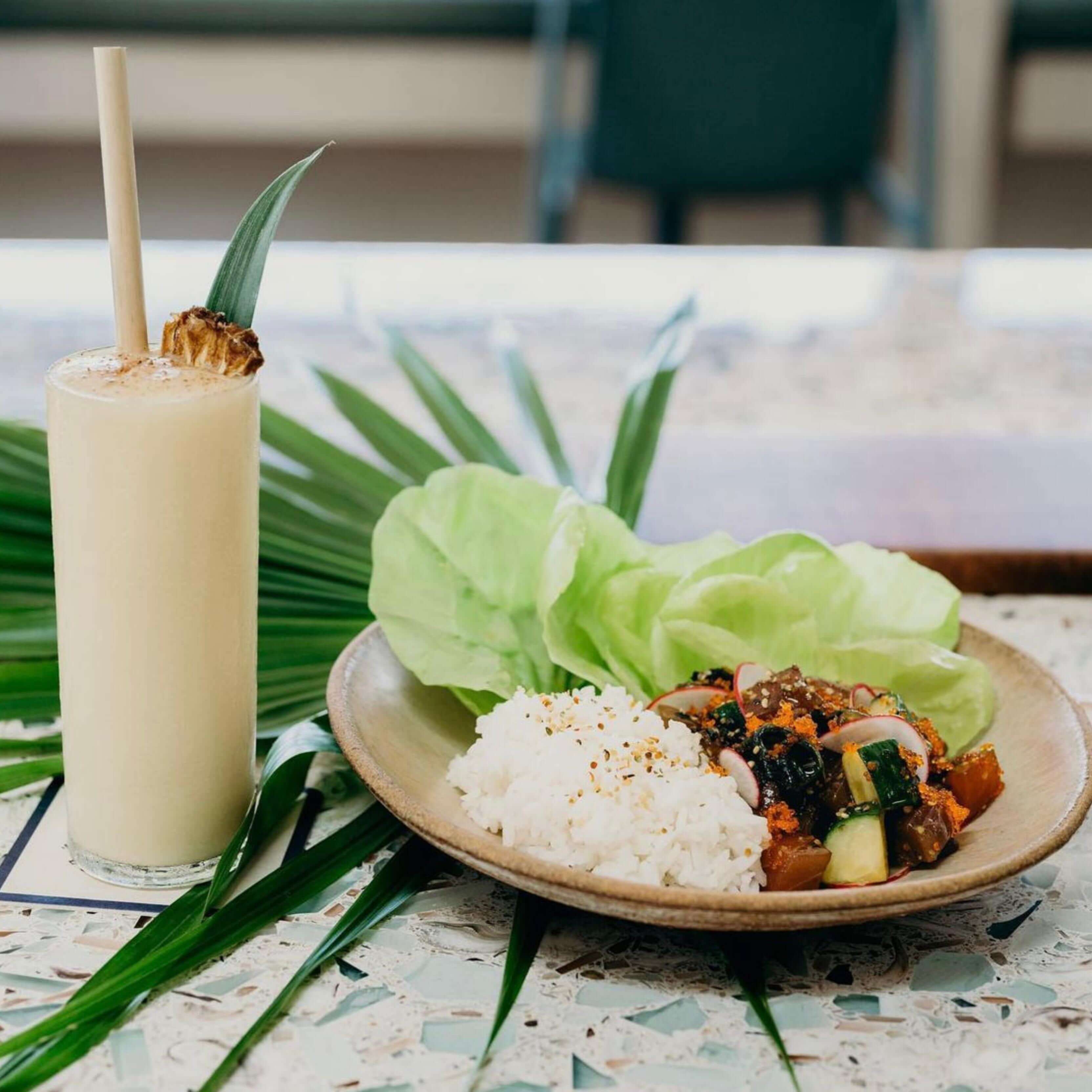 A piña colada garnished with a dried pineapple slice and a leaf is served in a tall glass with a bamboo-like straw made from reed stems. Next to it is a plate containing rice, lettuce leaves, and a mixed vegetable and seafood dish, all set on a patterned table with palm leaves. The arrangement creates a fresh and tropical atmosphere, emphasizing a sustainable and nature-inspired aesthetic.
